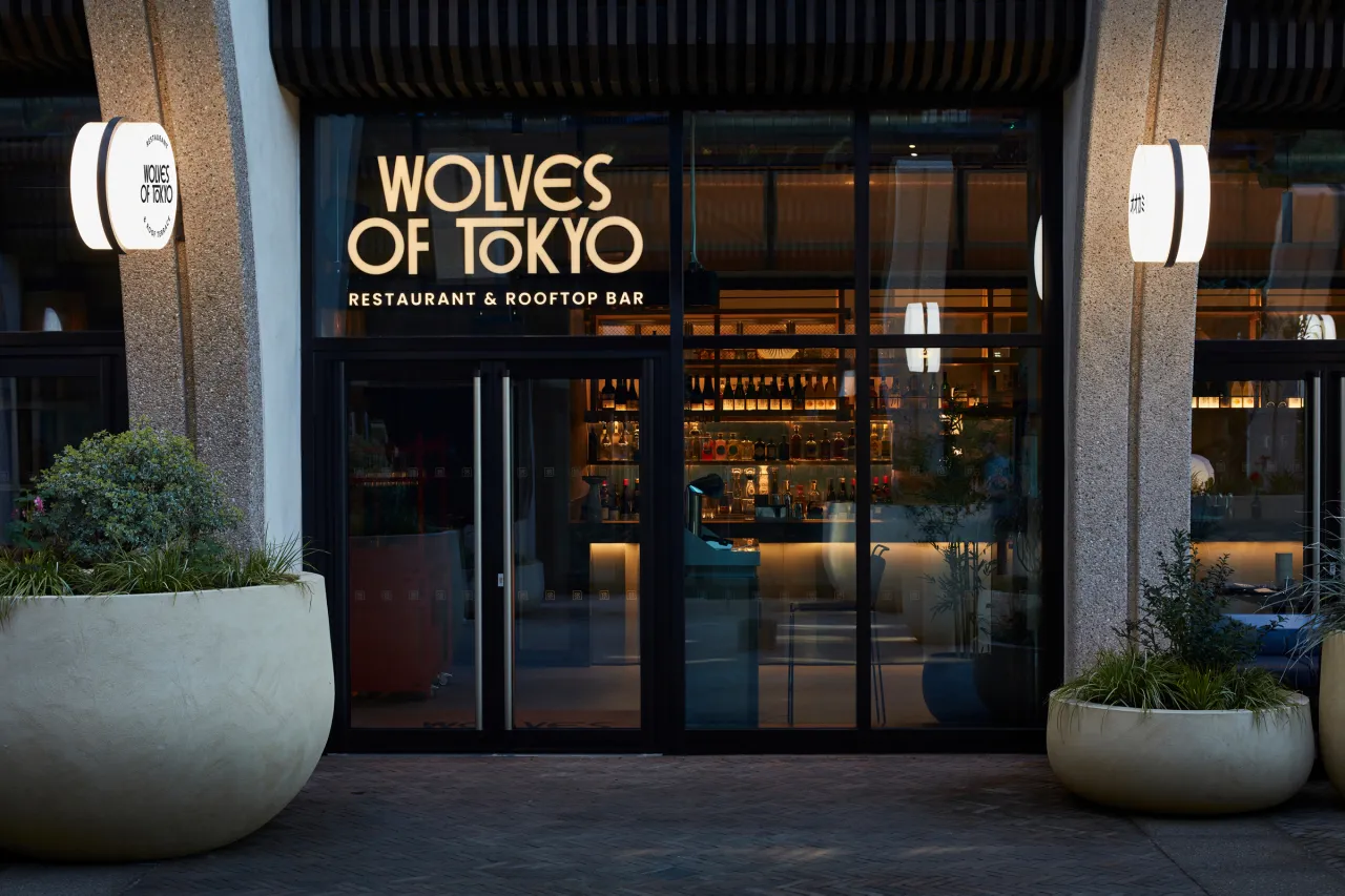 The illuminated storefront of Wolves of Tokyo restaurant and rooftop bar, featuring gold lettering on dark glass doors flanked by white wall lights and potted plants