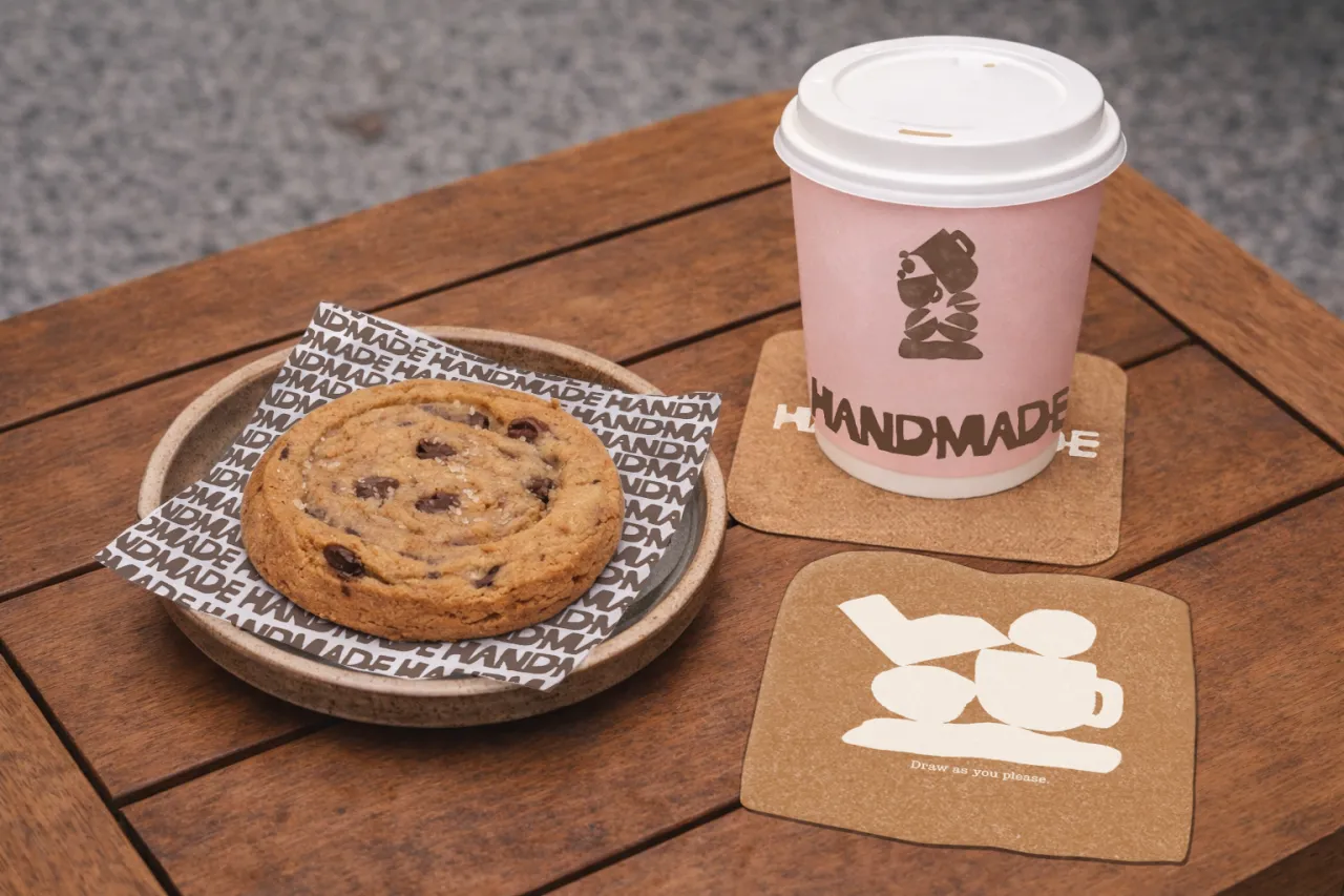 Handmade Café branding displayed on a wooden table with a chocolate chip cookie on a patterned plate, a pink takeaway cup and kraft paper coasters