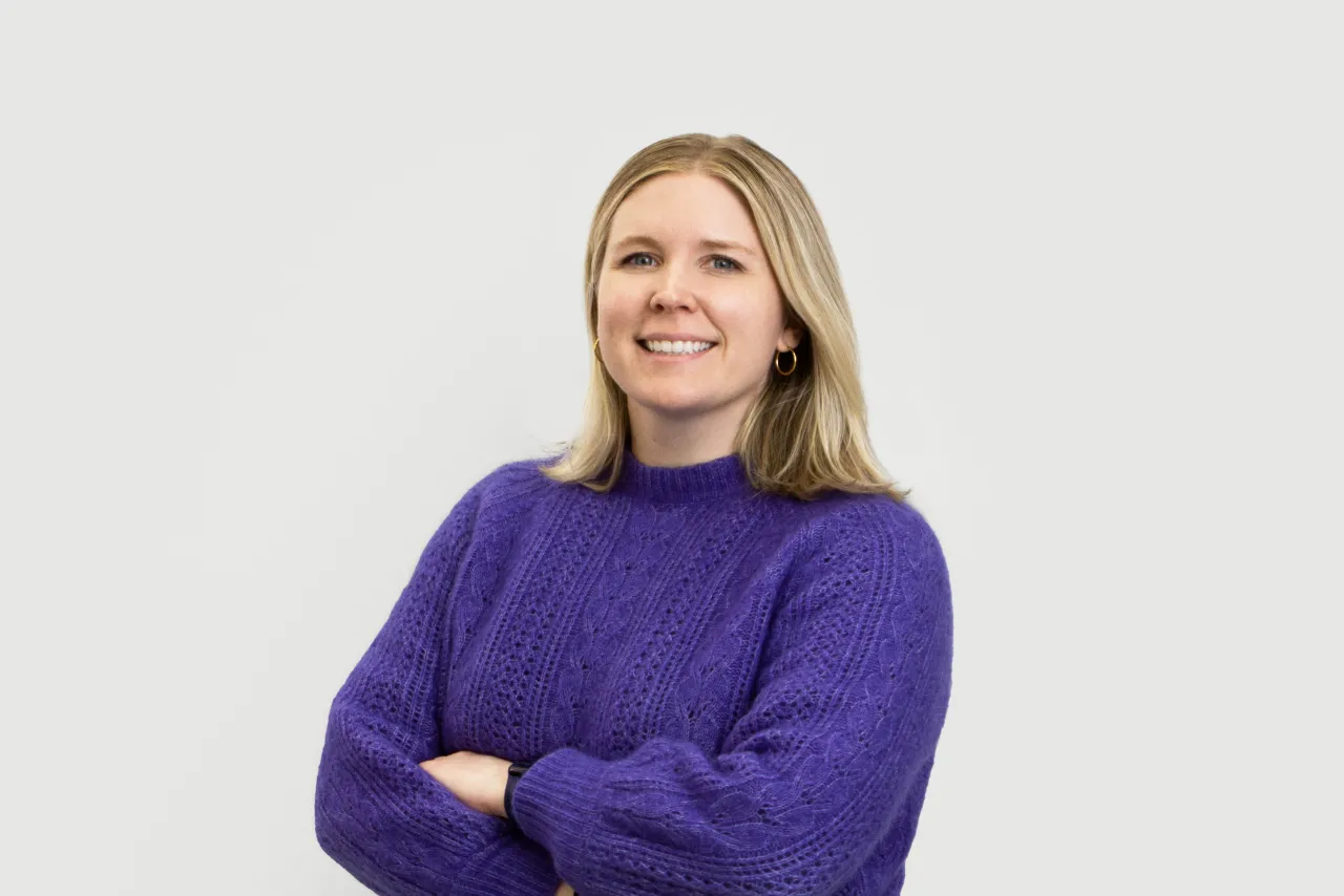 Woman with shoulder-length blonde hair wearing a purple cable-knit jumper, smiling at the camera with arms crossed against a white background