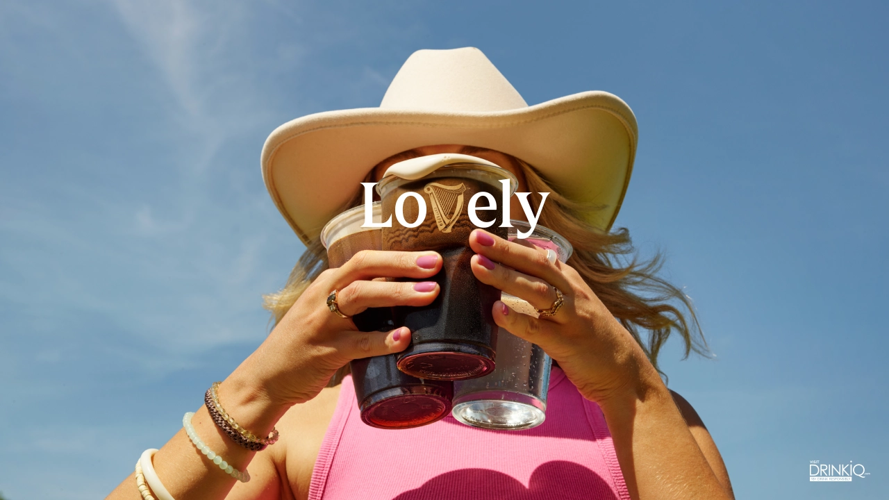 Person wearing a tan cowboy hat and pink top holds two glasses of Guinness against a blue sky, with the word "Lovely" displayed across the image